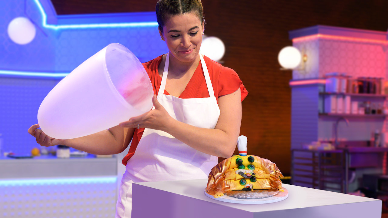 woman lifts a cloche to reveal a cake shaped like three croissants stacked on top of each other